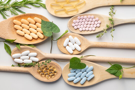 Wooden spoons holding various health supplements, soft gels, capsules, tablets, on white background.  Fresh green leaves, vines, and herb accents.