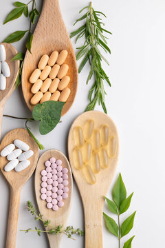 Wooden spoons holding various health supplements, soft gels, capsules, tablets, on white background.  Fresh green leaves, vines, and herb accents. vertical.