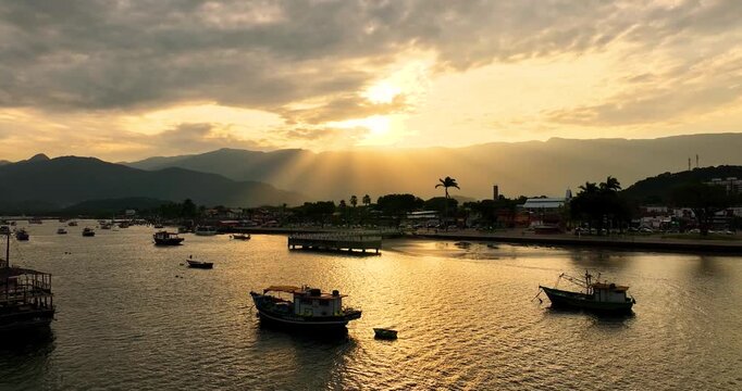 4K drone view of Bertioga waterfront at sunset, with fishing boats, golden reflections and Serra do Mar mountains in the background.