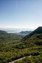 Fototapeta premium High angle panoramic view of Skadar lake valley in Montenegro with green forest trees on mountain hillsides and asphalt road asphalt curve line path in foreground and clear blue sky horizon background