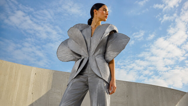 Futuristic woman in a sculptural silver couture outfit posing against modern concrete architecture and a cloudy blue sky.