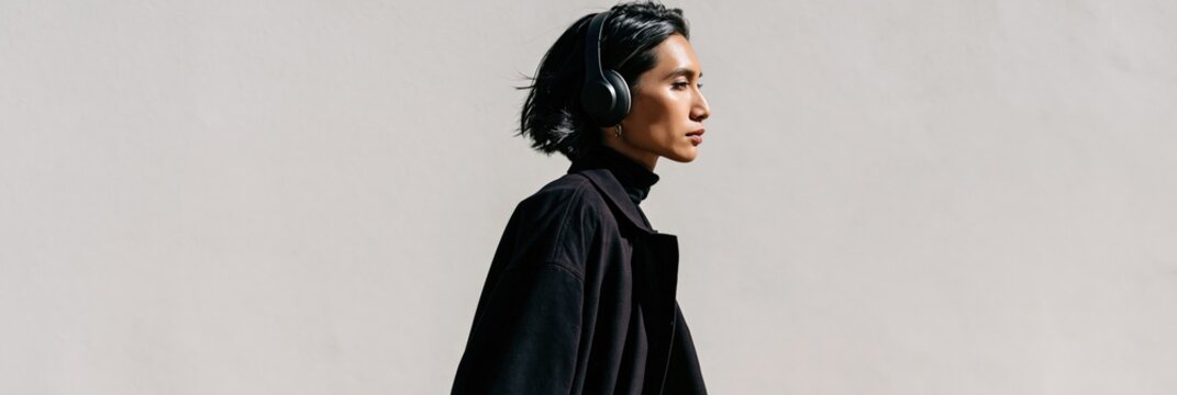 Side profile of a woman wearing headphones and a black coat walking by a pale wall for minimalist banner and urban lifestyle background design