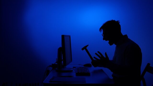 Man in silhouette experiencing extreme anger and frustration, holding a hammer over a computer monitor, shouting from intense stress and technology failure on a bright blue background