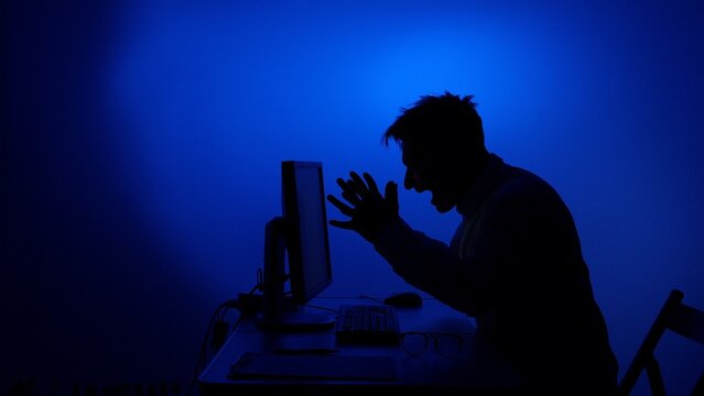 Man experiences digital frustration and anger while screaming at a desktop computer, his silhouette highlighted against a dramatic blue background, symbolizing stress