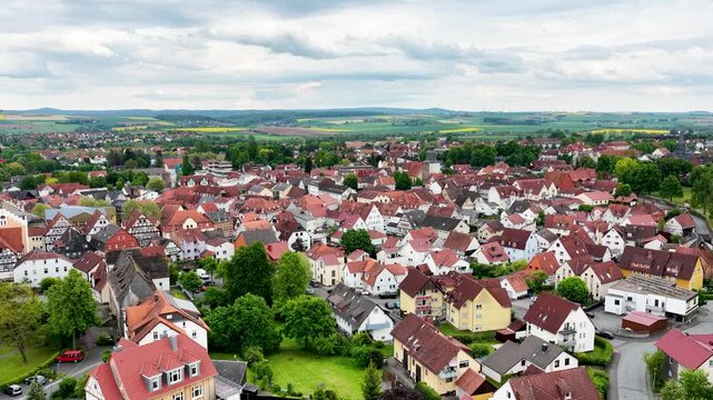 Blick &uuml;ber die D&auml;cher der historischen Altstadt von Hofgeismar mit der markanten Altst&auml;dter Kirche im Zentrum, umgeben von gr&uuml;nen B&auml;umen und sanften H&uuml;geln unter bew&ouml;lktem Himmel.