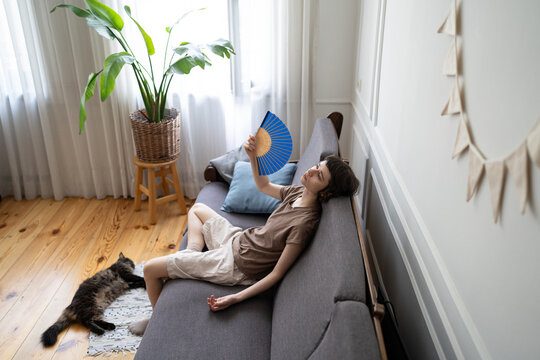 Exhausted woman fanning herself on sofa while cat lying nearby in heat distress. Indoor heat suffering, pet owner and animal under stress, summer heat without AC, discomfort, dehydration at home