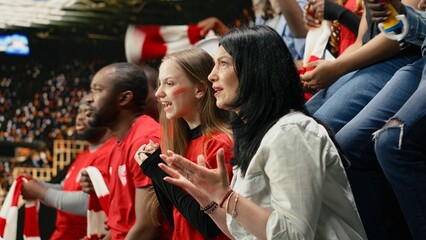 Obraz premium Excited female soccer fans leaning forward and cheering from stadium seats with diverse supporter crowd, red shirts and scarves during live match action.
