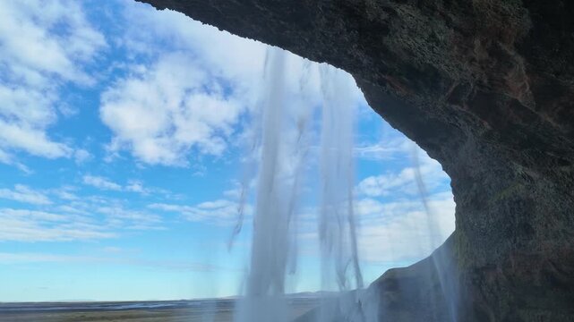 Vid&eacute;o film&eacute;e depuis l'int&eacute;rieur de la cavit&eacute; situ&eacute;e derri&egrave;re la cascade de Seljalandsfoss. Le rideau d'eau encadre le paysage de la c&ocirc;te sud islandaise sous un ciel bleu parsem&eacute; de nuages.