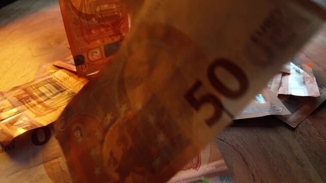 Plan rapproch&eacute; de billets en euros tombant au ralenti sur une table en bois. Les coupures se superposent progressivement tandis que la cam&eacute;ra capture les d&eacute;tails des billets et leur mouvement.