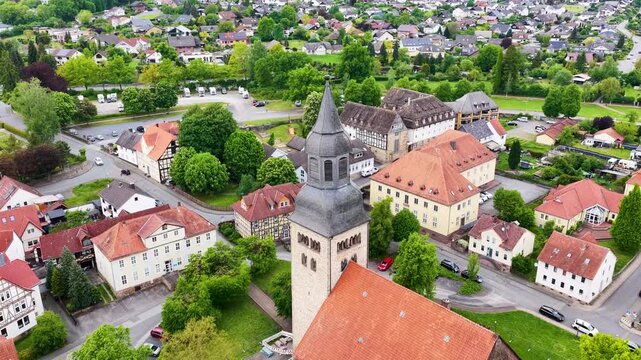 Blick &uuml;ber die D&auml;cher der historischen Altstadt von Hofgeismar mit der markanten Altst&auml;dter Kirche im Zentrum, umgeben von gr&uuml;nen B&auml;umen und sanften H&uuml;geln unter bew&ouml;lktem Himmel.