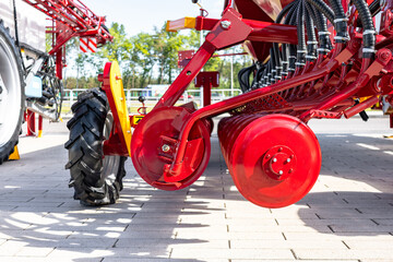 Low-angle view of a red seeder or planter machine parked on a paved outdoor yard. Visible elements include large black tractor tire, red metal discs and frames © NastyaPhoto
