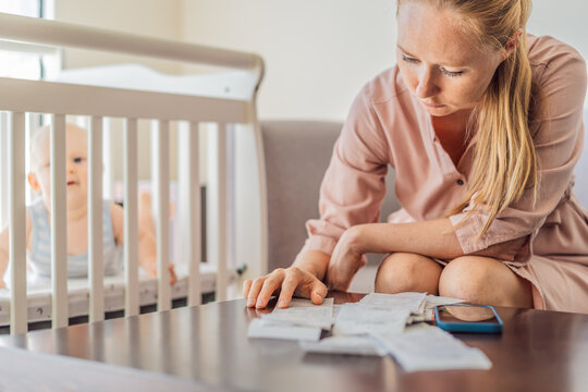 Mother sitting on sofa calculating monthly expenses with receipts while baby lies in crib nearby at home. Stressed mom managing budget and childcare. Financial pressure and parenthood concept