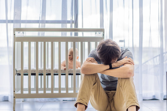 Tired father sitting by baby crib at home watching sleeping infant. Exhausted dad caring for newborn at night. Parenting fatigue, childcare responsibility, love and real family life concept