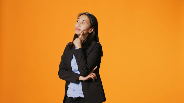 Thoughtful businesswoman posing over orange backdrop, thinking about new ideas and being in doubt, with clueless uncertain person brainstorming solutions, acting focused in front of camera.