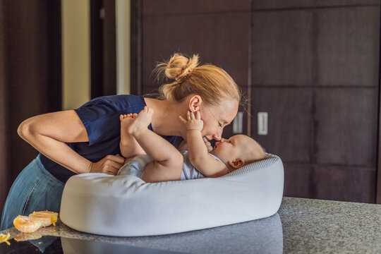 Mother talking to baby lying in cozy lounger on kitchen table while cooking at home. Caring mom interacting with infant during daily routine. Parenting, multitasking and family life concept