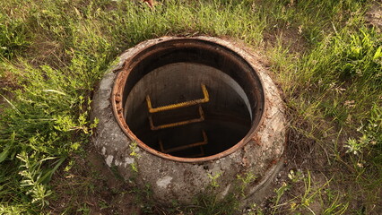 Otwarty betonowy właz kanalizacyjny z metalową drabiną w dół, otoczony zieloną trawą. © tomo81