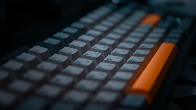 Close up macro shot of a mechanical computer keyboard with white, dark grey and orange keycaps in dark moody lighting.