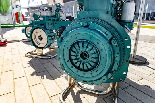 A large weathered cast-iron industrial pump painted turquoise with peeling paint sits on a paved plaza under bright sunlight. The circular pump housing