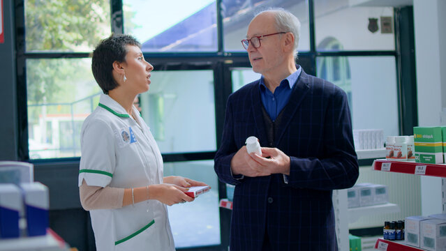 Aged man in pharmacy asking pharmacist for medical opinion, needing treatment for heart ache, with qualified pharmacist helping senior client choose best pharmaceutical cure for his disease.