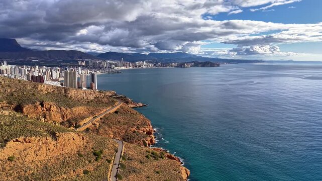 Stormfront Approach Over Coastal Road With Distant City Skyline, Churning Clouds, Deep Blue Sea And Rugged
