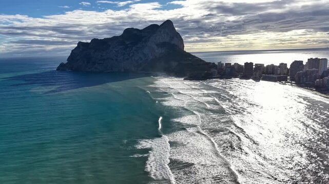 Rocky Headland Beside Shimmering Coastal City, Aerial Sweep Over Surf And Sandy Beach, Towering Rock