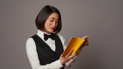 Naklejka premium Asian receptionist reading a literature book over grey background, enjoying lecture hobby with a novel story in studio, hotel concierge employee reads a fiction tale for knowledge.