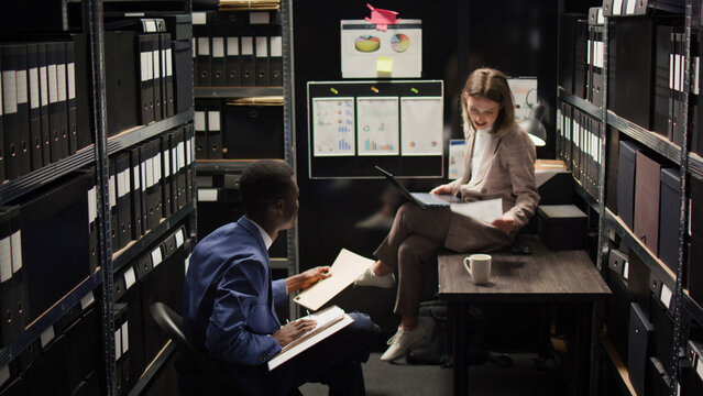 In room crammed with clues and records, Caucasian female investigator sits on table analyzing evidence with police officer, African American inspector hands over case data to policewoman with laptop.