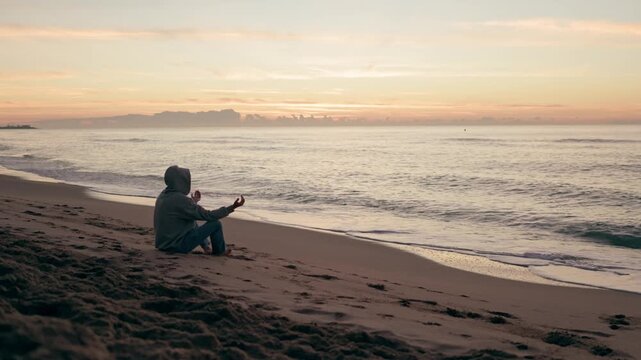 Young man wearing a hoodie practicing yoga and meditation on a sandy beach, sitting with his legs crossed while watching the ocean waves during a beautiful and peaceful sunrise