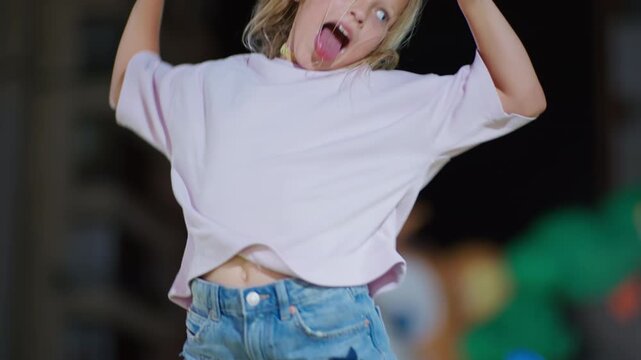 Young blonde girl making rock on hand gesture with both hand while jumping, sticking tongue out, smiling, filmed in slow motion
