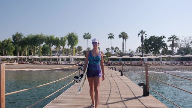 Rear view of a woman carrying wakeboard gear on a wooden pier by calm blue sea, with boats and islands ahead, creating a sunny summer mood for travel, leisure and water sport preparation themes.