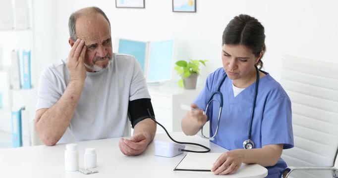 Nurse measuring man's blood pressure with sphygmomanometer and writing results in notebook at table in hospital