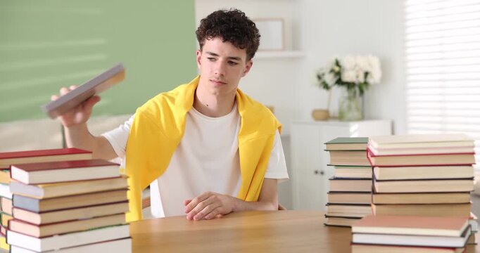Teenage boy reading and many books at wooden table indoors