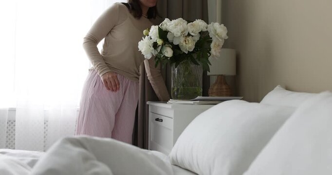 Woman putting vase with beautiful peonies on bedside table and turning on lamp at home, closeup