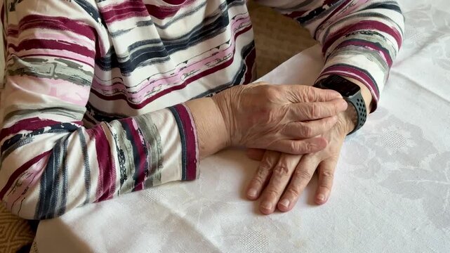 Close-up of an elderly woman's hands wearing a smartwatch. The device displays heart rate information. The woman has wrinkled skin and is wearing a patterned shirt