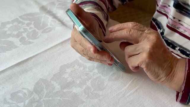 Close-up view of worn, wrinkled hands of an older person tapping and scrolling a touchscreen smartphone while seated at a white patterned tablecloth