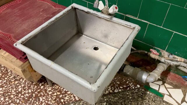 Rusty iron foot sink under faucet, set against green ceramic tiles, exposed pipes and valve, stained basin floor with clogged