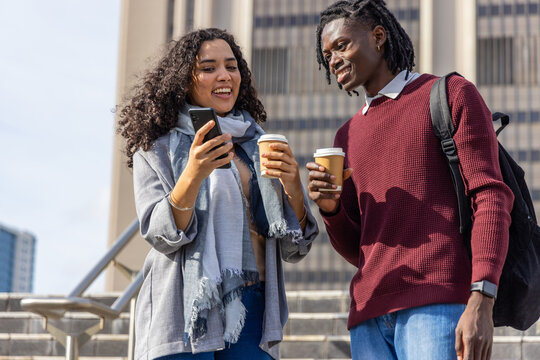 Casual couple chatting on stone steps on campus, woman holding smartphone, both holding cups
