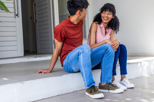 Asian man telling story to African American woman while sitting on stoop outside doorway smiling