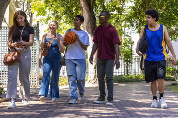 Naklejka premium Diverse friends walking along shaded brick path, carrying basketball, bags, wearing sporty clothes