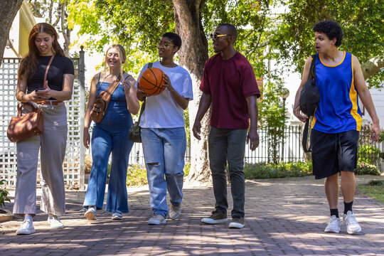 Diverse friends walking along shaded brick path, carrying basketball, bags, wearing sporty clothes