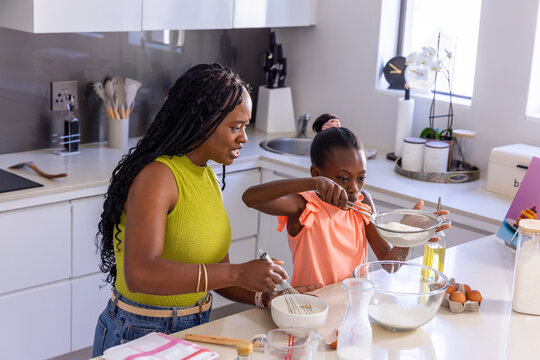 African American mother and teenage daughter baking in kitchen, sifting flour into glass bowl