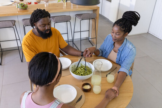 African American family holding hands and bowing heads at wooden table in kitchen with salad bowl