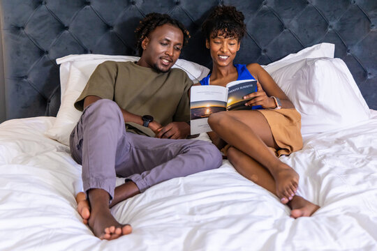 African American couple reclining on bed reading book with sunset picture, wearing wristbands