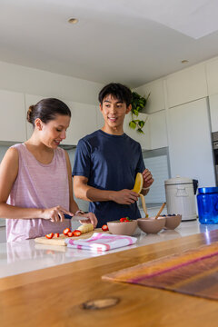 Diverse couple preparing fruit bowls at kitchen island, slicing strawberries and peeling banana