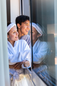 Diverse couple standing at doorway wearing white robes and terry headband reflecting in glass