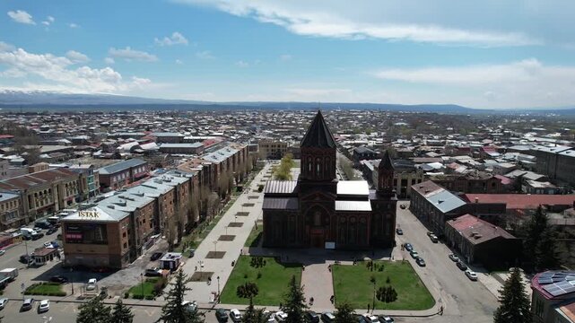 An aerial drone shot of the historic Holy Saviour Church in Gyumri, Armenia, showcasing its unique black tufa stone architecture and the surrounding cityscape on a sunny day.

