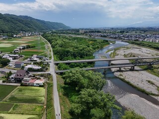 Obraz premium River flows calmly through rural landscape. Bridge spans water, connecting villages and fields. Green hills rise softly in the background. Road curves gently beside the river's edge