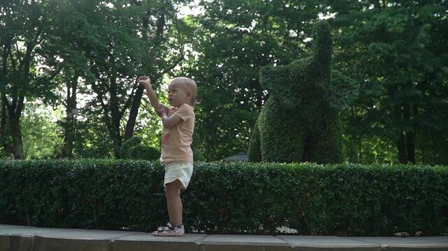 A small child, girl, points figure elephant from bush, park with landscaping. On your trunk and on your nose. Laughs, smiles, jumps. In shorts, T-shirt, sandals, with short hair. Day, summer