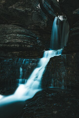 Dark rock walls frame a narrow waterfall. Water cascades softly over smooth stone. Long exposure blurs the moving water. Cool blue light adds serene depth. Ideal for nature, relaxation. © Thomas E. Lee