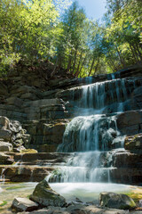 Naklejka premium Multi-tiered waterfall cascades over smooth, mossy rocks. Sunlight glints off flowing water at each level. Lush green forest surrounds the rocky stream. Bright daylight enhances natural textures